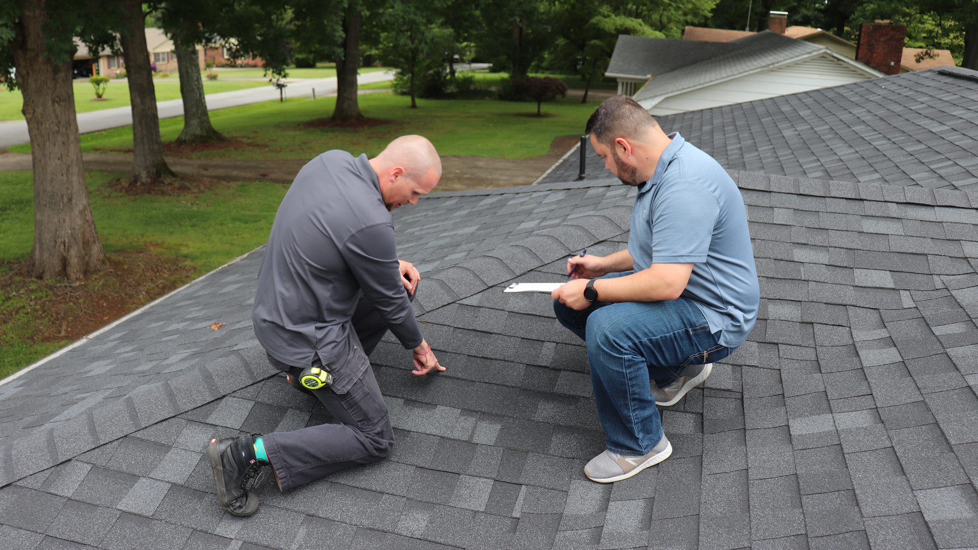 MBA Roofing inspectors kneel on a shingled roof after a storm, one pointing out a specific spot while the other documents it on a clipboard.